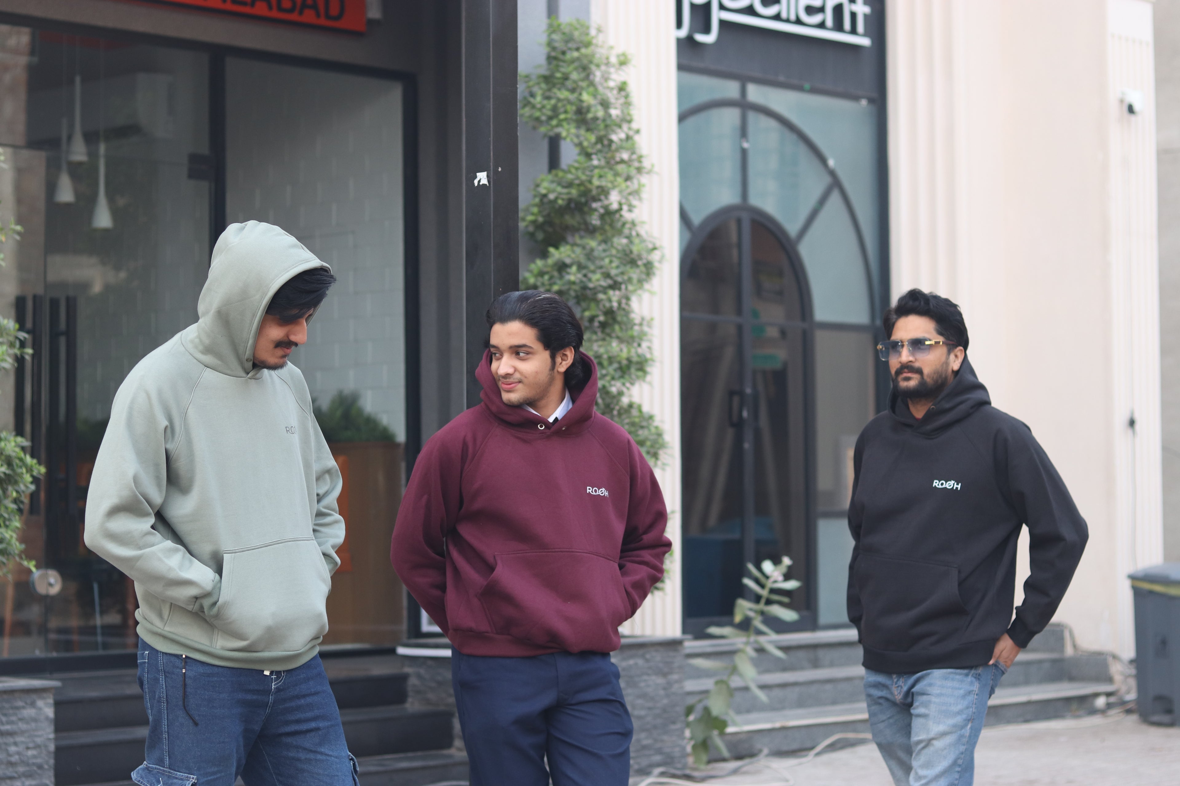 Three men wearing hoodies standing outside a building with a store sign in the background.
